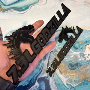 May include: Black 7.3L Godzilla emblem with a Godzilla head silhouette and the text "7.3L GODZILLA". The emblem has red eyes and is displayed on a blue and white background.