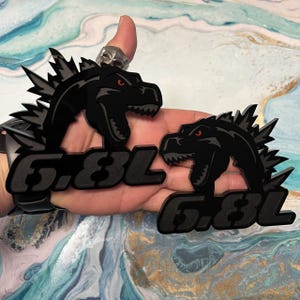 May include: Two black acrylic car emblems featuring a Godzilla-like monster with red eyes and the text "6.8L". The monster has a spiky dorsal fin and an open mouth. The emblems are likely for a vehicle.