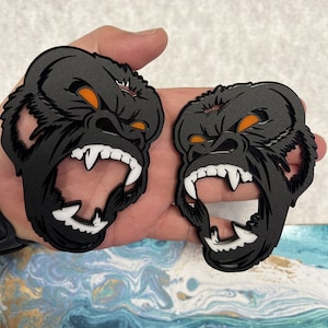 May include: Two black gorilla head-shaped metal ornaments with orange eyes and white teeth. The gorillas are shown with their mouths open in a roar.