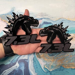 May include: Two black metal emblems featuring a Godzilla-like creature with red eyes and the text "7.3L". The emblems have a textured finish and are held in a hand.
