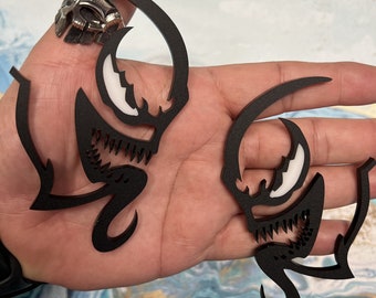 May include: Two black and white Venom symbiote earrings. The earrings are shaped like the Venom symbiote's head with a white outline and black teeth.