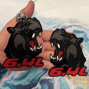 May include: Two black hippo-shaped emblems with open mouths, red tongues, and white teeth are displayed. The eyes are red. Below each hippo is the text "6.4L" in red. A silver ring is visible in the background.