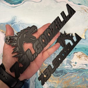 May include: Black Zilla Godzilla emblem with a carbon fiber pattern. The emblem features a Godzilla head with red eyes and the text "ZILLA GODZILLA". The emblem is held in a hand.