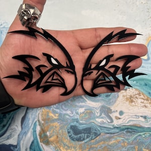 May include: Two black vinyl decals of stylized eagle heads with tribal-inspired designs. The eagle heads are facing each other and have white eyes.