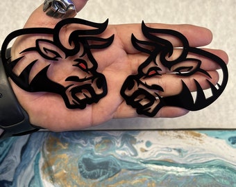 May include: Two black bull head silhouettes with red eyes. The bull heads are facing opposite directions and are cut out of a black material.