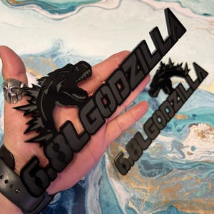 May include: Black Godzilla emblem with the text "6.8L GODZILLA" in a stylized font. The emblem features a detailed silhouette of Godzilla's head and dorsal fins. The emblem is held in a hand.