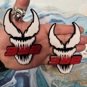 May include: Two white and black Venom-inspired car emblems with red 392 lettering. The emblems are held in a person's hand.
