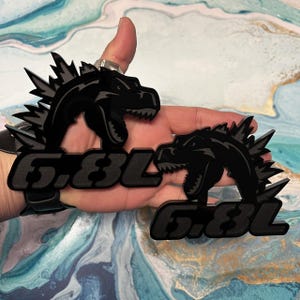 May include: Two black Godzilla-themed emblems with the text "6.8L". The emblems feature a detailed silhouette of Godzilla's head and dorsal fins. The emblems are likely for automotive customization.