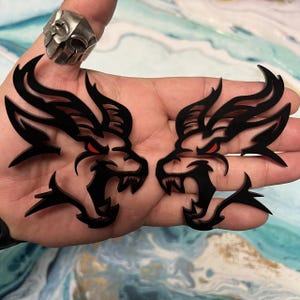 May include: Two black dragon head cutouts with red eyes and open mouths. The dragon heads are held in a hand. A silver skull ring is on the index finger. The background is a blue and white marbled surface.