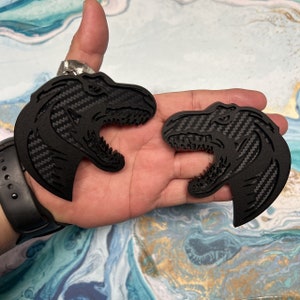 May include: Two black dinosaur-shaped keychains with a carbon fiber texture. The keychains are held in a hand.
