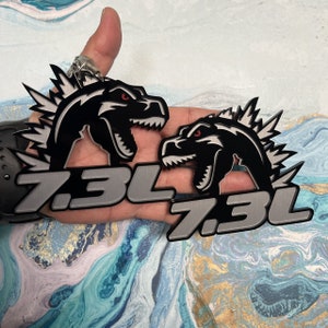 May include: Two black and silver metal car emblems with a dinosaur design. The emblems feature the text "7.3L" in silver with a black outline.