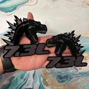 May include: Two black, stylized dinosaur emblems with the text "7.3L". The dinosaur has a spiky dorsal fin and an open mouth. The emblems are held in a hand against a colorful, abstract background.