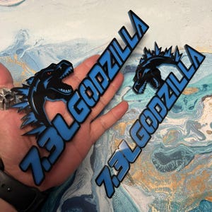 May include: Two black and blue Godzilla-themed decals. The decals feature the text "7.3L GODZILLA" in blue with a black outline, and a Godzilla graphic with red eyes. The decals are held in a hand.