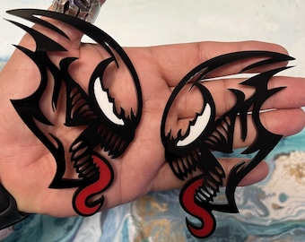 May include: Two black, stylized, symmetrical face designs with white eyes and red tongues. The designs have sharp, angular shapes and are held in a hand. The designs appear to be cut from a glossy material.