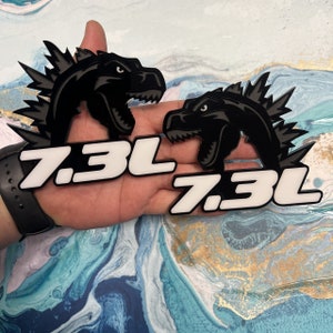 May include: Two black and white 7.3L decals with a dinosaur silhouette. The decals are on a hand.