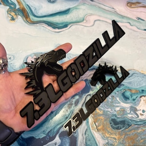 May include: Black metal emblem featuring the text "7.3L GODZILLA" with a Godzilla silhouette. The emblem is in a stylized font and has a glossy finish. The image shows multiple emblems.
