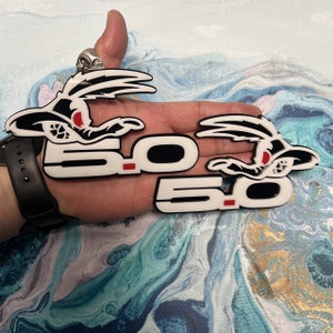 May include: Two white and black 5.0 emblems with a cartoon roadrunner graphic. The emblems have a red accent.