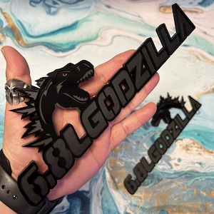 May include: Black Godzilla emblem with the text "6.8L GODZILLA" in a stylized font. The emblem features a Godzilla silhouette with red eyes. The image shows the emblem held in a hand.