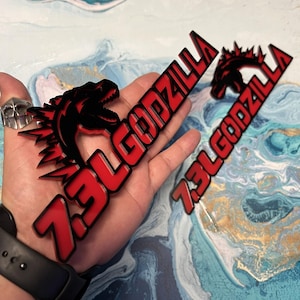 May include: A red and black Godzilla logo with the text "73L GODZILLA" in a stylized font. The logo features a silhouette of Godzilla's head and dorsal fins. The image is of a product.