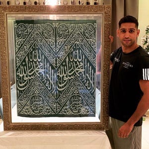 May include: A framed green textile with intricate white Arabic calligraphy. The frame is a light brown color. The textile is displayed on a white surface.