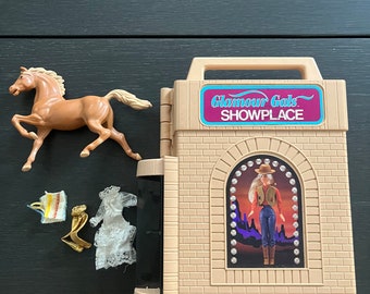 May include: A tan-colored Glamour Gals Showplace playset with a horse figurine, accessories, and a doll image. The playset features a handle and a doll image in a window. Accessories include a dress and a gold-colored item.