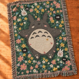 May include: A rectangular woven throw blanket featuring a dark green background with a pattern of colourful flowers and small white rabbit-like creatures. A large grey creature with a cream-coloured belly is centred, surrounded by black soot sprites.