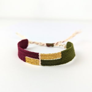 Può includere: Un braccialetto di amicizia intrecciato con fili color borgogna, oro e verde. Il braccialetto ha un semplice design a nodi ed è legato con un cordoncino bianco.