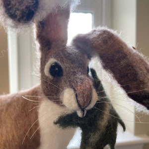 May include: A brown and white needle felted hare with long ears holding a green needle felted leaf in its mouth.