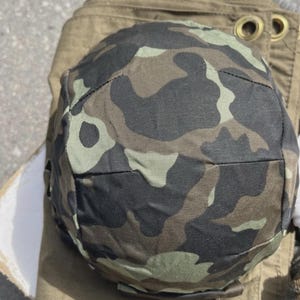 May include: A camouflage pattern helmet in shades of green, brown, and black. The helmet is resting on a khaki-coloured fabric. The helmet's design features a rounded shape with a slight brim.