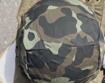 May include: A camouflage pattern helmet in shades of green, brown, and black. The helmet is resting on a khaki-colored fabric. The helmet's design features a rounded shape with a slight brim.