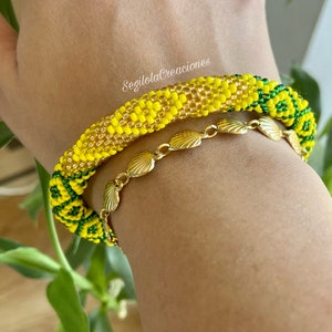 May include: A yellow and green beaded bracelet with a gold chain bracelet featuring shell-shaped links. The bracelet is worn on a person's wrist. The text "SegilolaCreaciones" is visible on the bracelet.