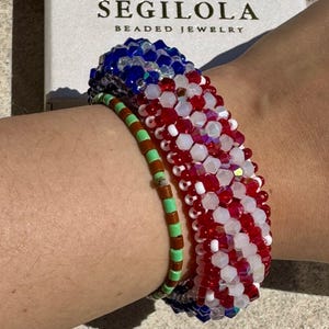May include: A wrist adorned with a beaded bracelet in red, white, and blue, reminiscent of the American flag. A smaller bracelet with brown and green beads is also present. The jewellery is from the brand Segilola.
