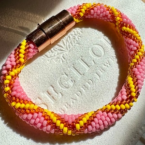 Puede incluir: Un brazalete de cuentas rosa y amarillo con un cierre magnético dorado. El brazalete está hecho de pequeñas cuentas redondas y tiene un patrón tejido.