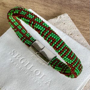 Könnte beinhalten: Ein Armband aus grünen und braunen Perlen mit sich wiederholendem Muster. Das Armband hat einen silberfarbenen Magnetverschluss. Das Armband liegt auf einer weißen Oberfläche mit dem geprägten Text "SEGILOLA BEADED JEWELRY".