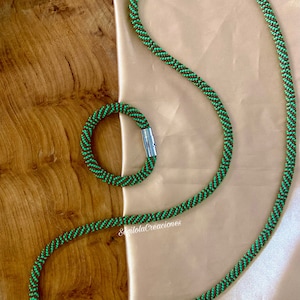 May include: A green and brown beaded necklace and bracelet set with magnetic clasps. The necklace is longer than the bracelet. The jewellery is displayed on a wooden surface.