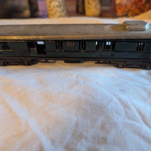Puede incluir: Modelo de vagón de tren antiguo. El vagón es de color verde oscuro con un techo gris y presenta varias ventanas y el número "354" en el lateral. El vagón está sobre una superficie blanca. Mide unos 20 cm de largo.