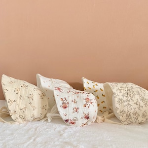 May include: Five baby bonnets in various floral and fruit patterns. The bonnets are made of soft, crinkled fabric and feature tie closures. Colors include cream, white, and shades of pink, yellow, and brown. The bonnets are arranged on a white surface.