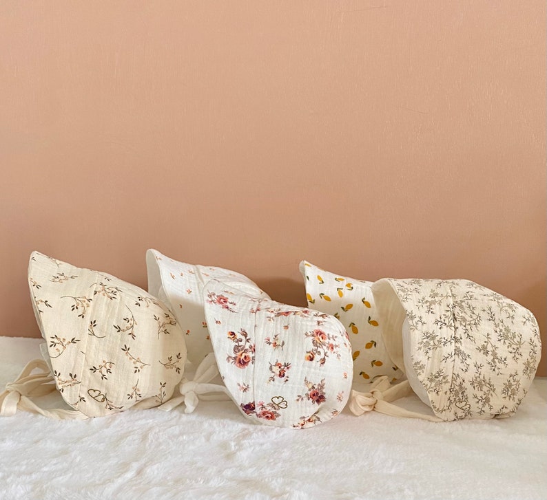 May include: Five baby bonnets in various floral and fruit patterns. The bonnets are made of soft, crinkled fabric and feature tie closures. Colors include cream, white, and shades of pink, yellow, and brown. The bonnets are arranged on a white surface.