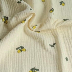 May include: Cream-colored fabric with a repeating pattern of yellow lemons and green leaves. The fabric has a textured, crinkled appearance, suggesting a soft, breathable material. Ideal for crafting or sewing projects.
