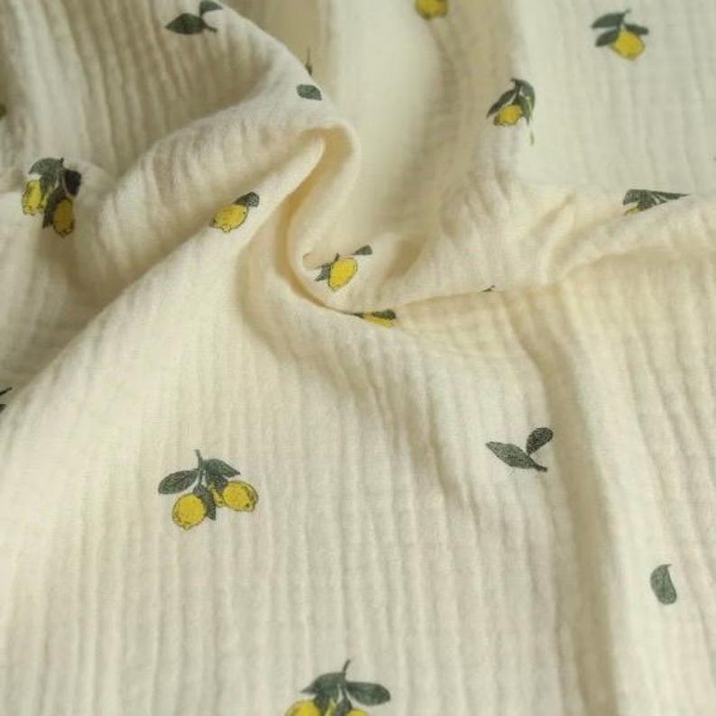May include: Cream-colored fabric with a repeating pattern of yellow lemons and green leaves. The fabric has a textured, crinkled appearance, suggesting a soft, breathable material. Ideal for crafting or sewing projects.