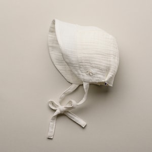 May include: A white baby bonnet made of a textured fabric, featuring a tie closure and a small gold embroidered cloud design. The bonnet is designed to protect the baby's head and face. The bonnet is a soft, neutral color.
