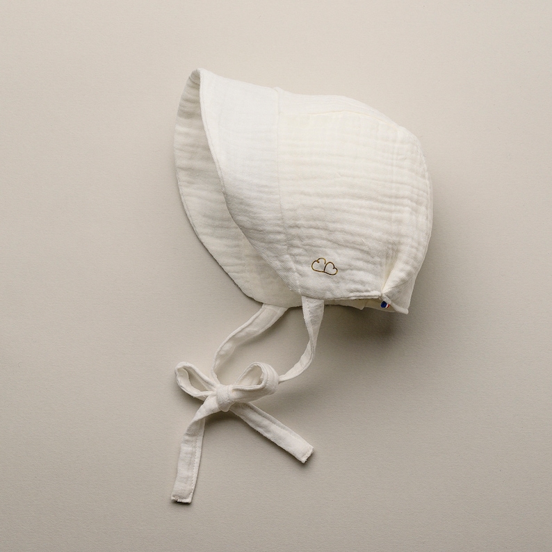 May include: A white baby bonnet made of a textured fabric, featuring a tie closure and a small gold embroidered cloud design. The bonnet is designed to protect the baby's head and face. The bonnet is a soft, neutral color.