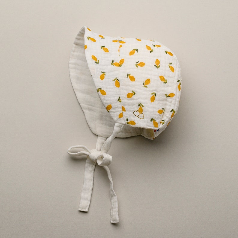 May include: White baby bonnet with a lemon pattern. The bonnet is made of a soft, textured fabric and features a tie closure under the chin. The lemon pattern includes yellow lemons with green leaves scattered across the white background.