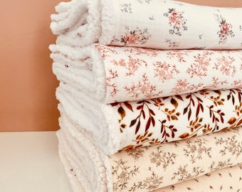 May include: A stack of five floral-patterned blankets. The top blanket has a white, fluffy trim and a cream background with pink flowers. The other blankets have various floral designs in shades of brown, cream, and purple. The blankets are stacked on a white surface.