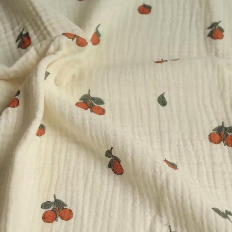 May include: Cream-colored fabric with a repeating pattern of orange fruit and green leaves. The fabric has a textured, crinkled appearance, suggesting a soft, lightweight material. The fruit design is stylized, with a vintage or handmade aesthetic.