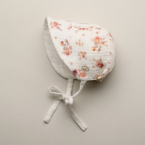May include: A white baby bonnet with a floral pattern in shades of peach, orange, and brown. The bonnet has a rounded shape with a brim and ties under the chin. The fabric appears to be a soft, lightweight material.