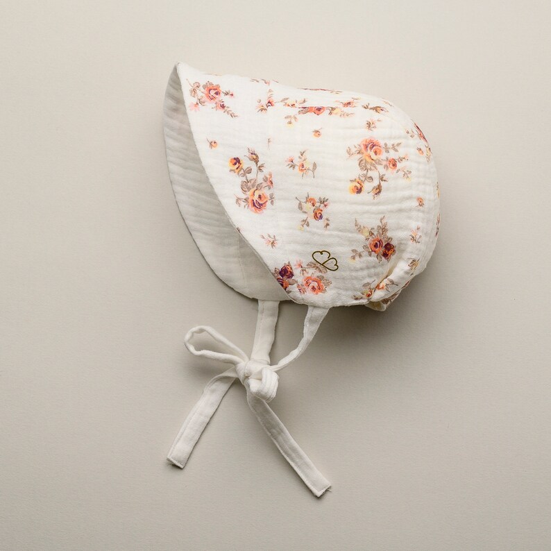 May include: A white baby bonnet with a floral pattern in shades of peach, orange, and brown. The bonnet has a rounded shape with a brim and ties under the chin. The fabric appears to be a soft, lightweight material.