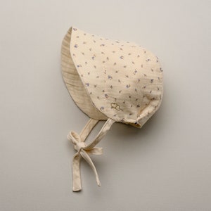 May include: A cream-colored baby bonnet with a floral pattern of small blue flowers. The bonnet has a soft, layered design with a tie closure under the chin. A small embroidered cloud logo is visible on the front.