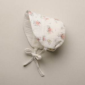 May include: A white baby bonnet with a floral pattern of pink roses and green leaves. The bonnet has a tie closure and a double-layered design. The fabric appears to be a soft, textured material.