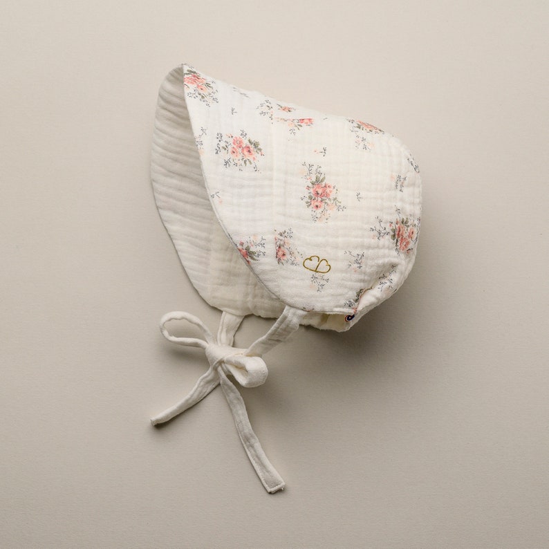 May include: A white baby bonnet with a floral pattern of pink roses and green leaves. The bonnet has a tie closure and a double-layered design. The fabric appears to be a soft, textured material.
